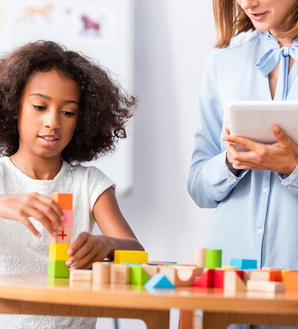 A young child builds with blocks as her occupational therapist looks on